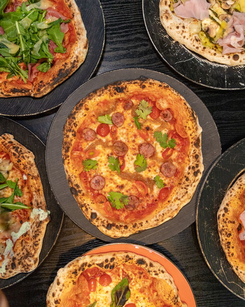 Cinco pizzas de masa madre recién horneadas vistas desde arriba sobre una mesa negra: margherita, jamón con aceitunas, espinaca con queso y prosciutto con rúcula